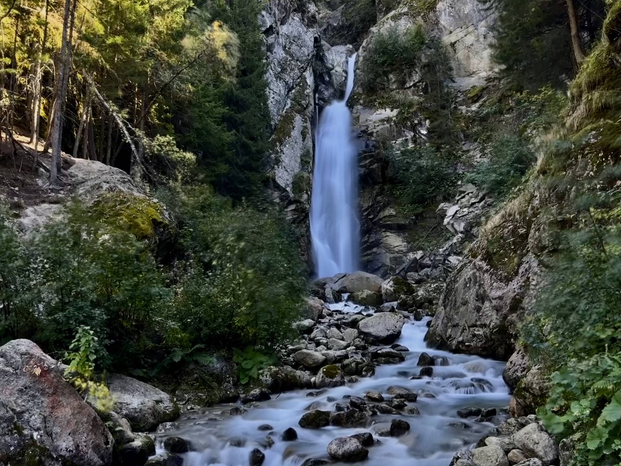 A milky waterfall tumbles down a cliff side with ghostly waters flowing through a creek at Cascade du Dard in Chamonix, France.