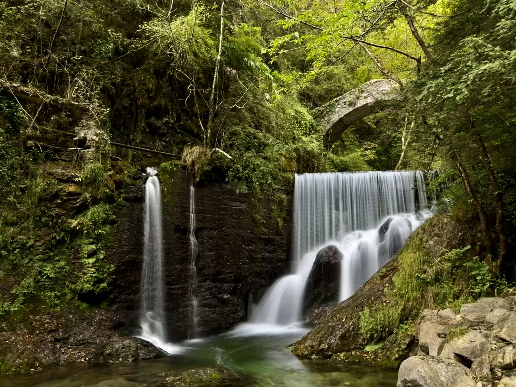 A waterfall plunges into a lush forest with a stone footbridge that crosses overhead near Lake Como, Italy.