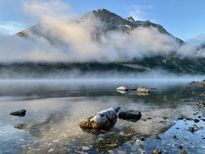 Colorful rocks in the lake with morning fog surrounding Red Eagle Mountain at Saint Mary Lake